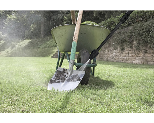 Un nettoyeur haute pression nettoie une pelle sale devant une brouette verte sur une pelouse.
