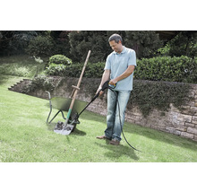 Un homme nettoie une pelle sale dans une brouette a l aide d un nettoyeur haute pression sur une pelouse dans un jardin.