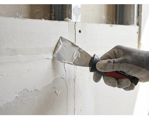 Hand mit Arbeitshandschuh trägt mit einem Rautner Malerspachtel Spachtelmasse auf ein Bewehrungsband an einer Trockenbauwand auf.