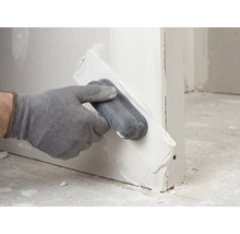 Hand mit Handschuh trägt Spachtelmasse mit einem Applikator auf eine Trockenbauwand auf.
