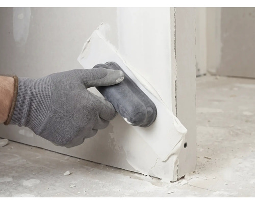 Hand mit Handschuh trägt Spachtelmasse mit einem Applikator auf eine Trockenbauwand auf.