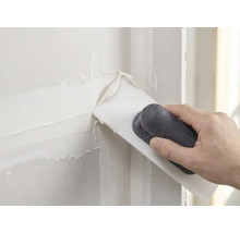 Hand trägt weiße Spachtelmasse mit einem Spachtel auf eine Wand auf.