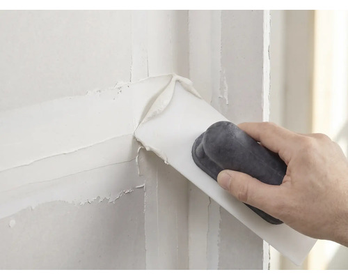 Hand trägt weiße Spachtelmasse mit einem Spachtel auf eine Wand auf.