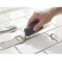 Eine Hand trägt mit einem Fugenspachtel graue Fugenmasse auf weiße Fliesen auf.