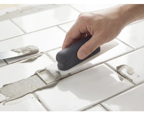 Eine Hand trägt mit einem Fugenspachtel graue Fugenmasse auf weiße Fliesen auf.