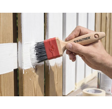 Hand streicht einen Holzzaun mit einem Rautner Flachpinsel und weißer Farbe.