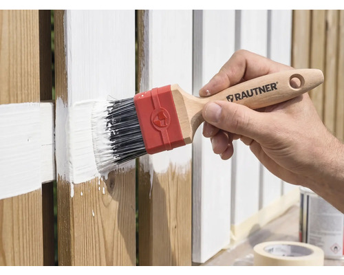 Hand streicht einen Holzzaun mit einem Rautner Flachpinsel und weißer Farbe.