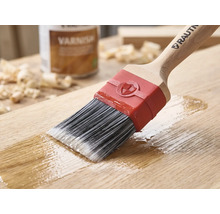 RAUTNER Pinsel trägt Klarlack auf eine Holzoberfläche auf. Mit Synthetikborsten, Holzgriff und roter Zwinge. Im Hintergrund eine Dose Lack.