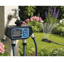 Digitale Bewässerungssteuerung von for.q an einem Außenwasserhahn mit angeschlossenen Gartenschläuchen und aktivem Rasensprenger im Garten.
