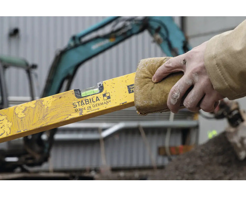 Une main nettoie un niveau à bulle avec Stabila Logo avec une éponge sur un chantier devant une pelleteuse.