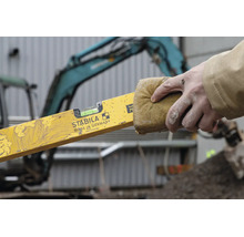 Une main nettoie un niveau à bulle avec Stabila Logo avec une éponge sur un chantier devant une pelleteuse.
