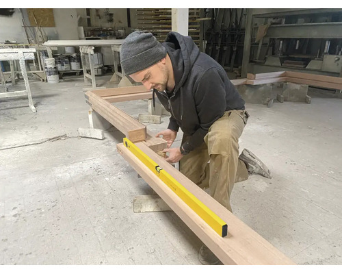 Un artisan agenouillé dans un atelier vérifie l'alignement d'un grand cadre en bois avec un niveau à bulle jaune.