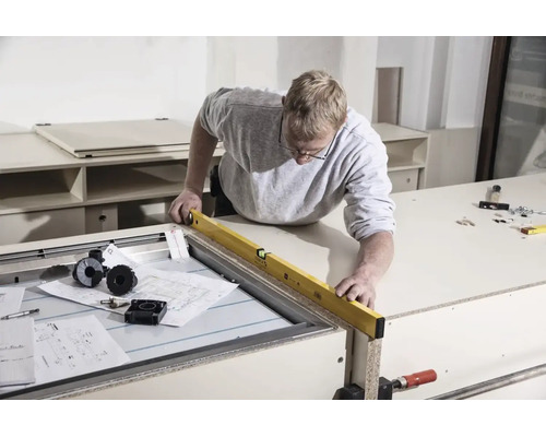 Un homme aligne un meuble à l'aide d'un niveau à bulle dans un atelier lumineux avec des outils et des plans de montage.