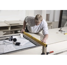 Un homme aligne un meuble à l'aide d'un niveau à bulle dans un atelier lumineux avec des outils et des plans de montage.