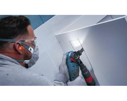 Artisan portant des lunettes de protection et un masque respiratoire sciant une découpe dans un panneau blanc avec un outil multifonction