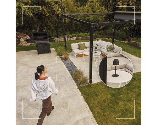 Salon de jardin moderne avec pergola noire, canapé d'angle, barbecue à gaz et une petite lampe de table noire sur une table basse.
