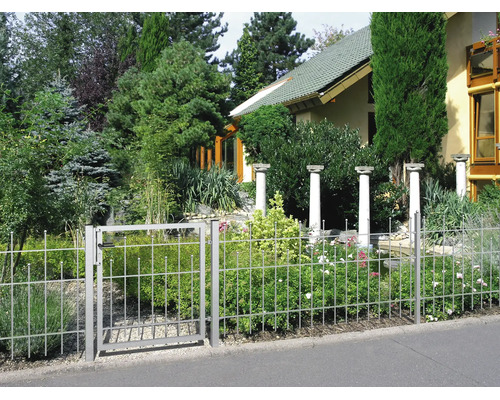 Metallzaun mit Gartentor in einem bepflanzten Garten mit Büschen, Bäumen und dekorativen Steinsäulen im Hintergrund.