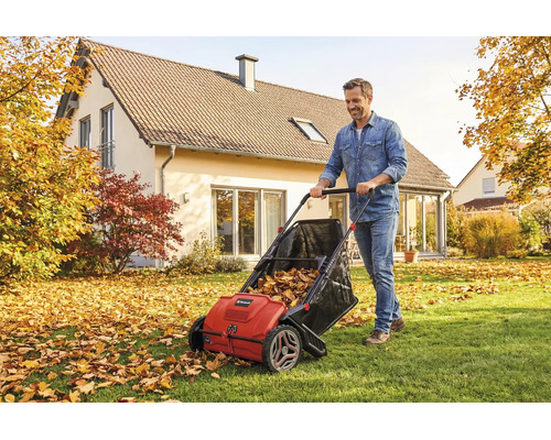 Un homme pousse un ramasseur de feuilles et d herbe rouge sur une pelouse parsemée de feuilles d automne devant une maison dans un cadre lumineux.