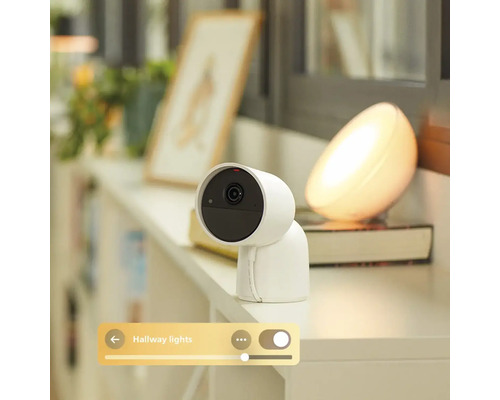 Caméra de surveillance intelligente blanche pour intérieur sur étagère, incluant le contrôle via une application de la lumière connectée.