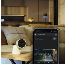Caméra de sécurité Philips Hue Secure sur une table à côté d'un smartphone affichant la vue en direct de l'application Philips Hue dans un salon.