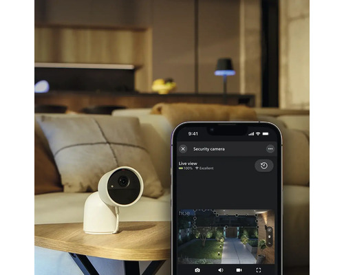 Caméra de sécurité Philips Hue Secure sur une table à côté d'un smartphone affichant la vue en direct de l'application Philips Hue dans un salon.