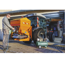 Un homme entretient un accessoire de tracteur à l'aide d'un compresseur Metabo et d'un pistolet de pulvérisation devant un garage.