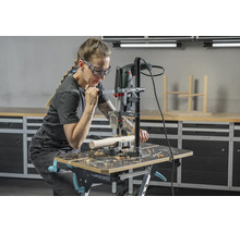 Personne utilisant un support de perceuse avec perceuse sur un établi dans un atelier lumineux, avec lunettes et protection auditive.