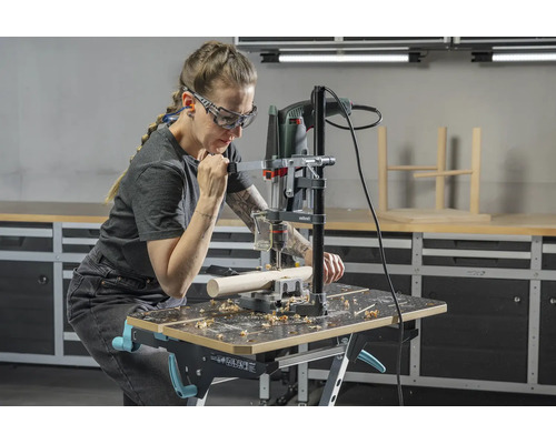 Personne utilisant un support de perceuse avec perceuse sur un établi dans un atelier lumineux, avec lunettes et protection auditive.