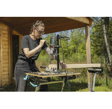 Femme utilisant un support de perçage et une perceuse pour percer une poutre en bois sur un établi mobile dans un jardin.