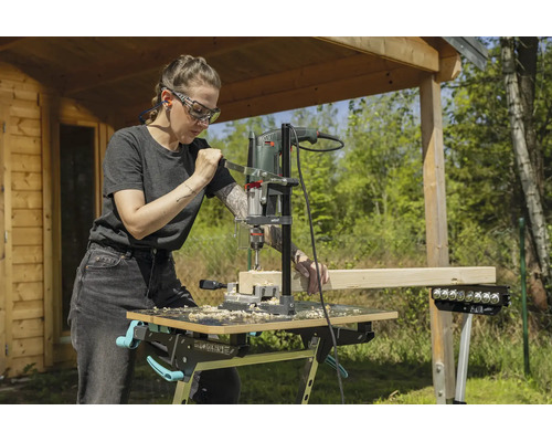 Femme utilisant un support de perçage et une perceuse pour percer une poutre en bois sur un établi mobile dans un jardin.