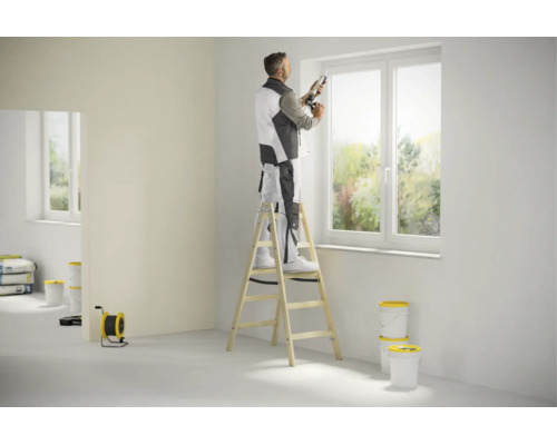 Un artisan sur un escabeau en bois scelle une fenêtre avec un pistolet à cartouche dans une pièce lumineuse.