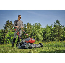Ein Mann mäht einen grünen Rasen mit einem Einhell Akku-Rasenmäher in einer sonnigen Gartenlandschaft mit Bäumen im Hintergrund.