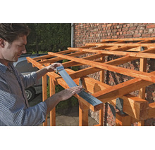 Ein Mann bringt ein silbernes Bitumenband an den Holzbalken einer Dachkonstruktion vor einer Ziegelwand an.