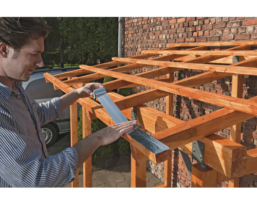 Ein Mann bringt ein silbernes Bitumenband an den Holzbalken einer Dachkonstruktion vor einer Ziegelwand an.