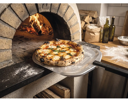 Pizza fraîchement cuite sur une pelle à pizza avec Tenneker Logo devant un four à pierre enflammé dans une cuisine rustique.