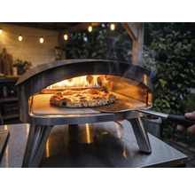 Four à pizza extérieur en acier inoxydable avec des flammes et une pizza sur pierre à pizza manipulée avec une pelle à pizza.