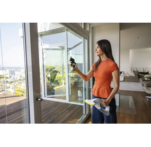 Une femme nettoie une grande porte vitrée de balcon avec un nettoyeur de vitres sans fil dans un salon lumineux et moderne.