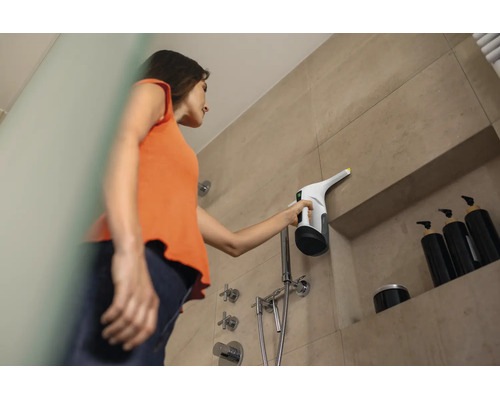 Une personne nettoie de grands carreaux de salle de bains dans la douche avec un nettoyeur de surfaces portatif dans une salle de bains moderne.