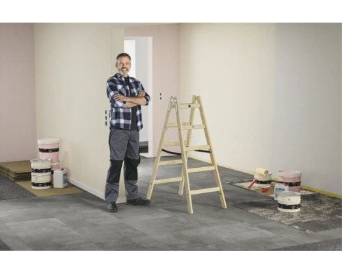 Handwerker steht neben einer Holz-Stehleiter in einem hellen, aufgeräumten Raum bei Renovationsarbeiten mit Farbkesseln und Malerwerkzeug am Boden.