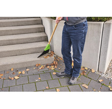 Une personne balaie des feuilles sèches sur un escalier en béton et un sol pavé à l'aide d'un balai à manche en bois et poils synthétiques.