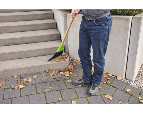 Une personne balaie des feuilles sèches sur un escalier en béton et un sol pavé à l'aide d'un balai à manche en bois et poils synthétiques.