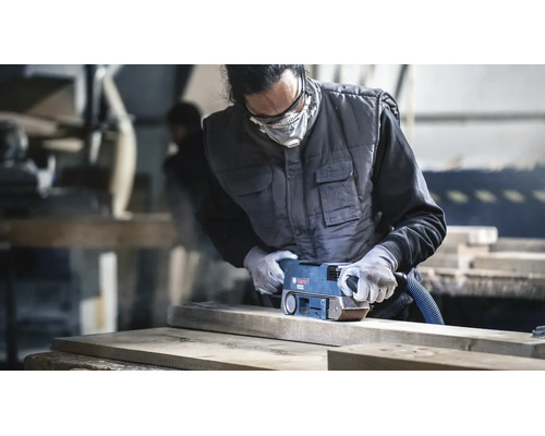Handwerker schleift ein Holzbrett mit einem Bosch Bandschleifer in einer Werkstatt mit Schutzkleidung und Atemschutzmaske.