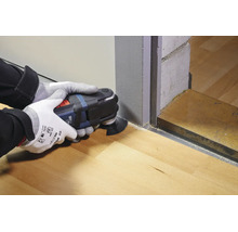 Personne avec des gants de travail utilisant un outil multifonction oscillant pour couper un cadre de porte en bois au ras du sol.