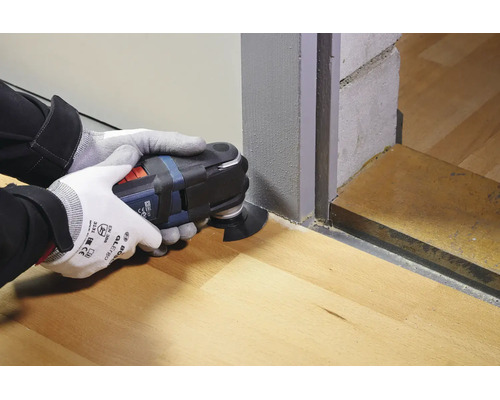 Personne avec des gants de travail utilisant un outil multifonction oscillant pour couper un cadre de porte en bois au ras du sol.