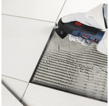 Un outil multifonction Bosch est utilisé pour enlever la colle à carrelage à côté d un carreau posé.