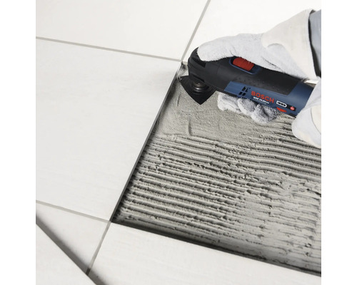 Un outil multifonction Bosch est utilisé pour enlever la colle à carrelage à côté d un carreau posé.