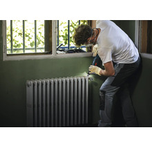 Artisan avec équipement de protection ponçant un vieux radiateur à éléments devant une fenêtre.
