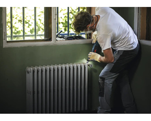 Artisan avec équipement de protection ponçant un vieux radiateur à éléments devant une fenêtre.
