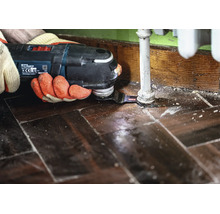 Un artisan utilise un outil multifonction Bosch avec lame de scie pour découper précisément le parquet autour d'un tuyau de chauffage.
