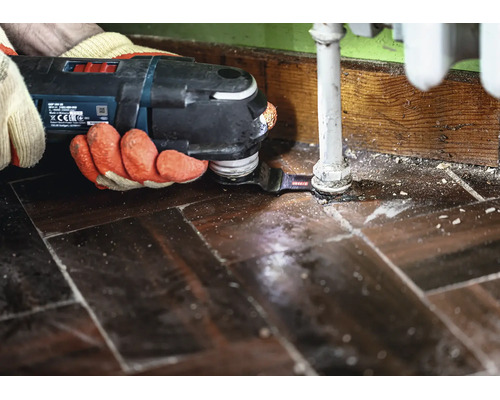Un artisan utilise un outil multifonction Bosch avec lame de scie pour découper précisément le parquet autour d'un tuyau de chauffage.